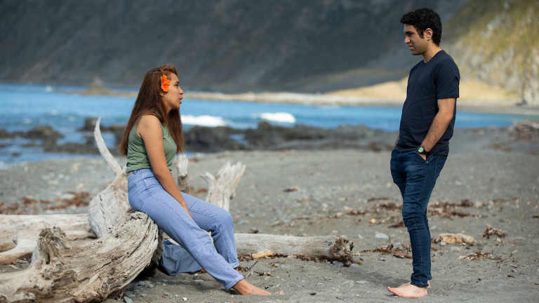 Young woman sitting on a log at the beach talking to a young man standing, with the sea behind them.